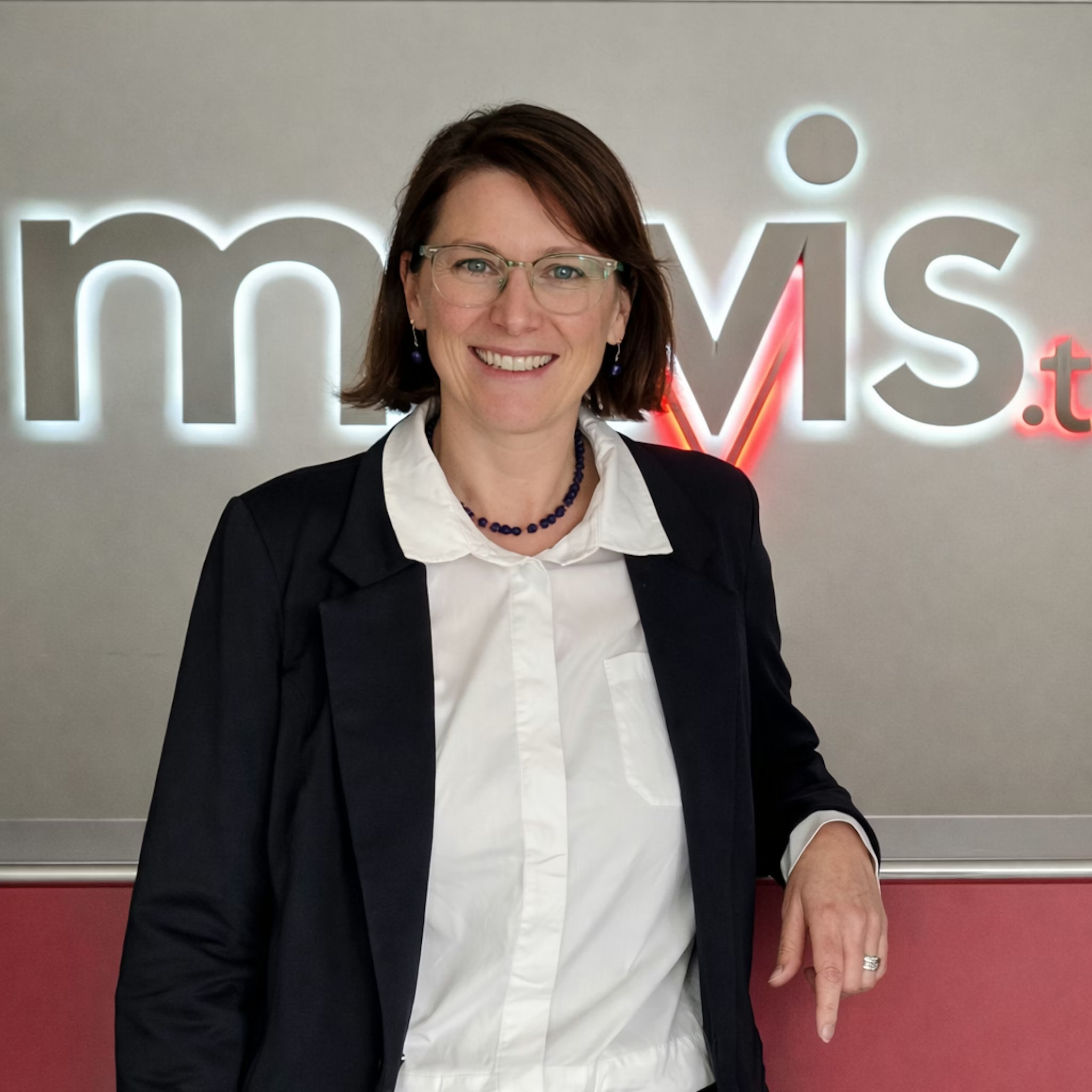Frau mit Brille, schwarzem Blazer, weißer Bluse und dunkler Jeans steht lächelnd vor einer Wand mit teilweise sichtbarem Logo.